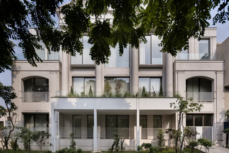 Street facade with white balconies, planted terraces and arched openings framed by overhanging tree branches