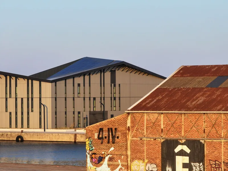 Waterside elevation of the beige paneled facade with vertical fenestration next to weathered brick warehouses at dusk