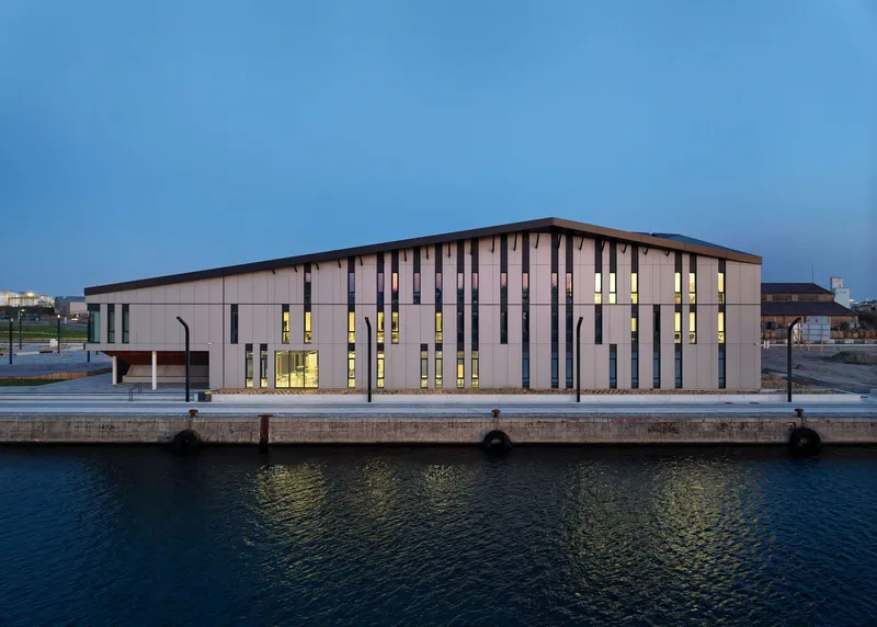 Dockside elevation showing vertical window openings illuminated between timber cladding panels at blue hour