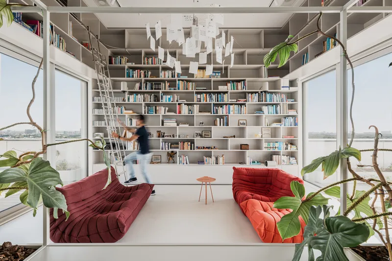 Floor-to-ceiling bookshelf wall with rolling ladder and a person in motion amid hanging plants