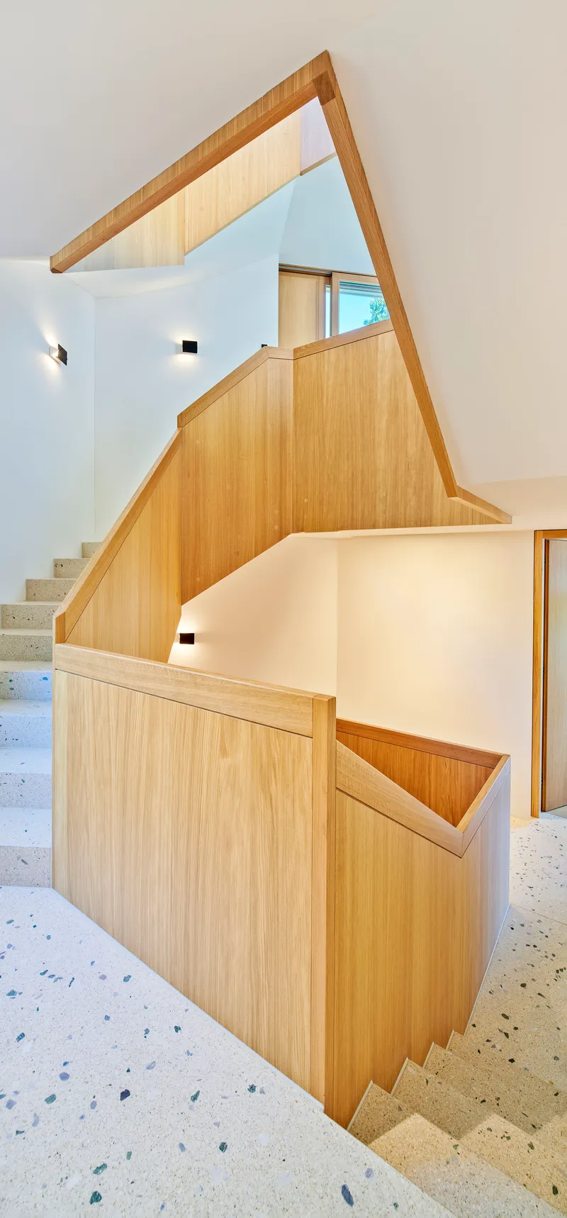 Angular timber-clad stair volume with terrazzo treads connecting multiple levels under a skylight