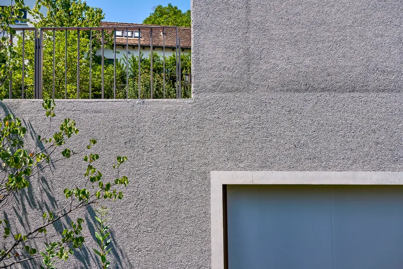 Close-up of textured concrete wall panels with metal railing and young tree casting shadows in sunlight
