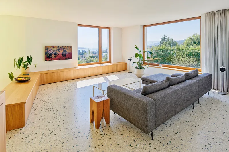 Living room with terrazzo flooring and timber-framed windows overlooking treetops in afternoon light