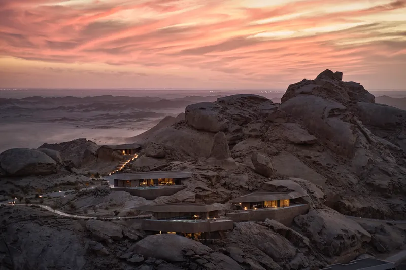 Terraced volumes nestled among boulder formations with illuminated interiors at dusk