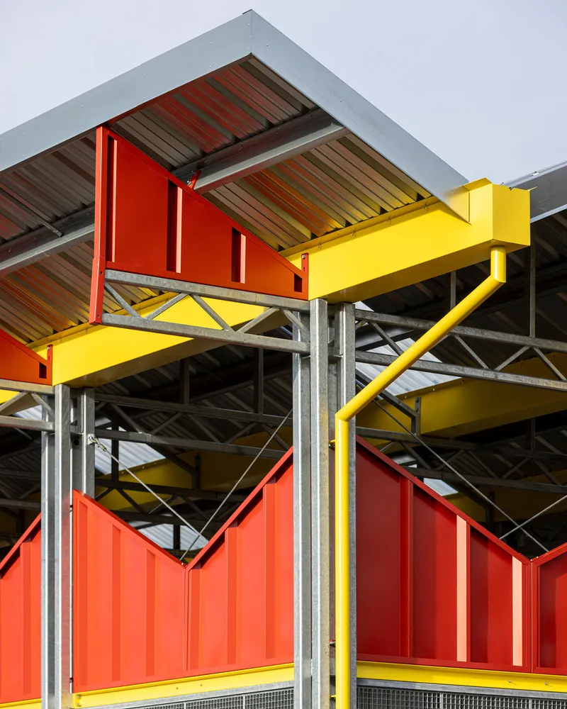Exposed steel truss beneath a corrugated metal roof supported by red and yellow structural columns