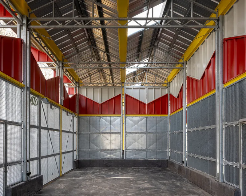 Interior corner showing mesh walls, red chevron panels, and exposed steel truss ceiling with skylights