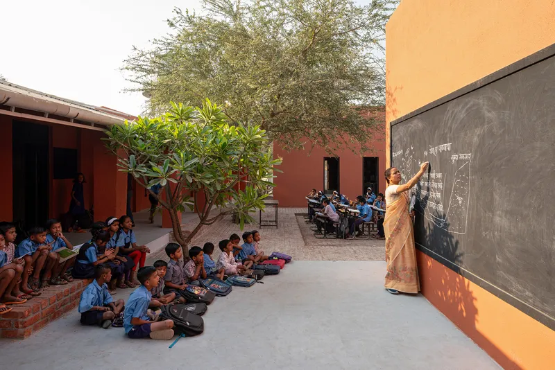 Open-air courtyard classroom with chalkboard mounted on ochre wall and students seated beneath a tree
