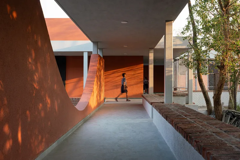 Covered walkway with curving terracotta wall and brick seating as children walk past under dappled afternoon light