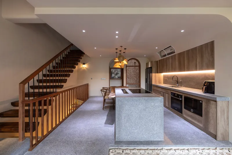 Open kitchen with terrazzo island, timber cabinetry, and vertical-slat stair balustrade under recessed lighting
