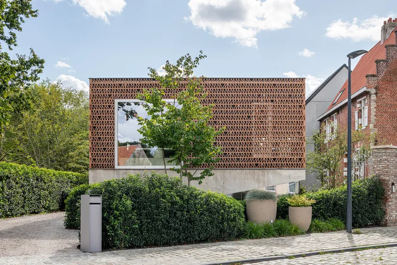 Street view of perforated terracotta brick facade raised on concrete plinth with hedge in foreground