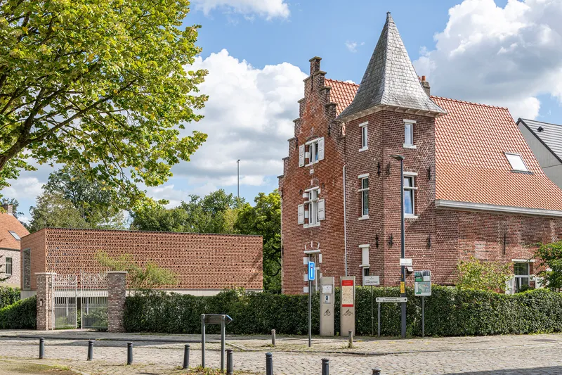 Low perforated brick addition adjacent to historic turret building with cobbled street and trees