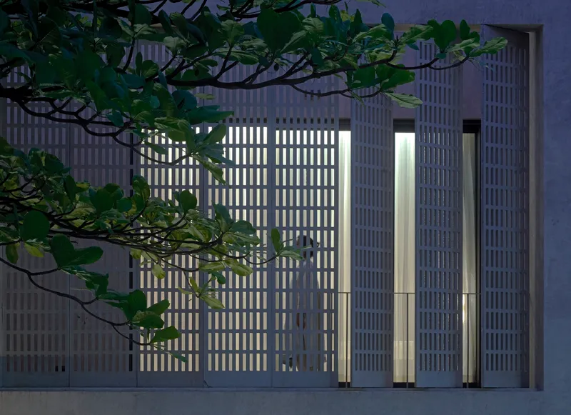 Illuminated vertical-slat facade at dusk with tree branches crossing the perforated concrete screen