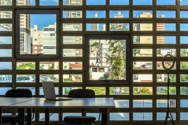 Interior view through concrete grid screen framing a table with laptop overlooking neighboring buildings and trees