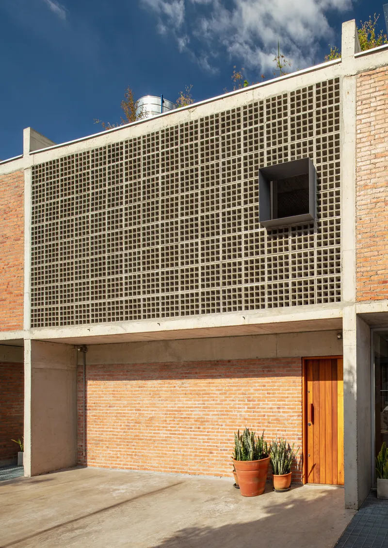 Entry courtyard with perforated concrete screen wall above timber door and potted plants under blue sky