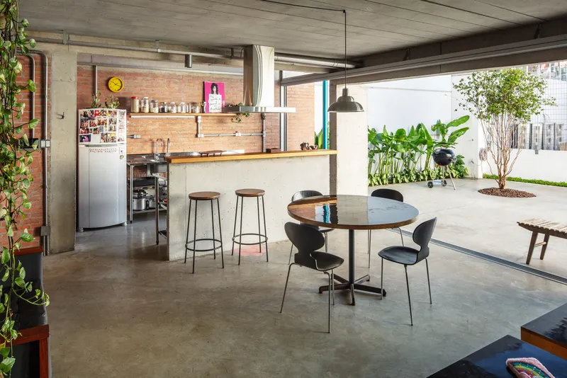 Open-plan interior with exposed concrete ceiling, kitchen counter, dining table and courtyard visible beyond