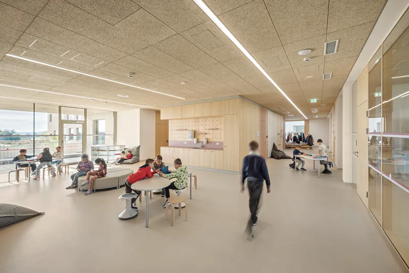 Interior corridor with house-shaped plywood structure and children at low tables under linear ceiling lights