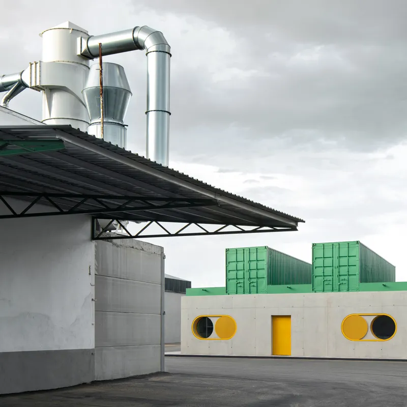 Concrete facade with circular yellow openings seen beyond an industrial shed with metal ventilation ducts