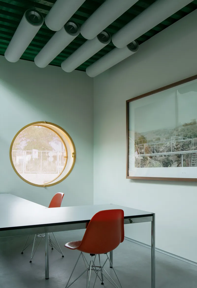 Meeting room with circular brass-rimmed window, red chairs at a white table, and exposed ceiling ducts