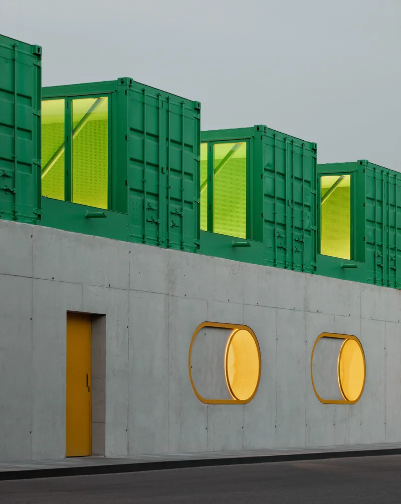 Concrete wall with two circular brass-rimmed windows and yellow door below three green shipping containers with yellow mesh panels