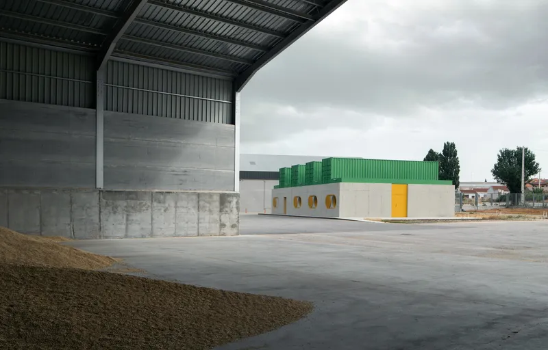 View from covered storage area showing poured concrete wall and green corrugated metal building across yard