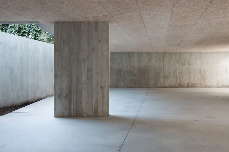 Ground level parking area with board-formed concrete columns and ceiling beneath a planted courtyard opening