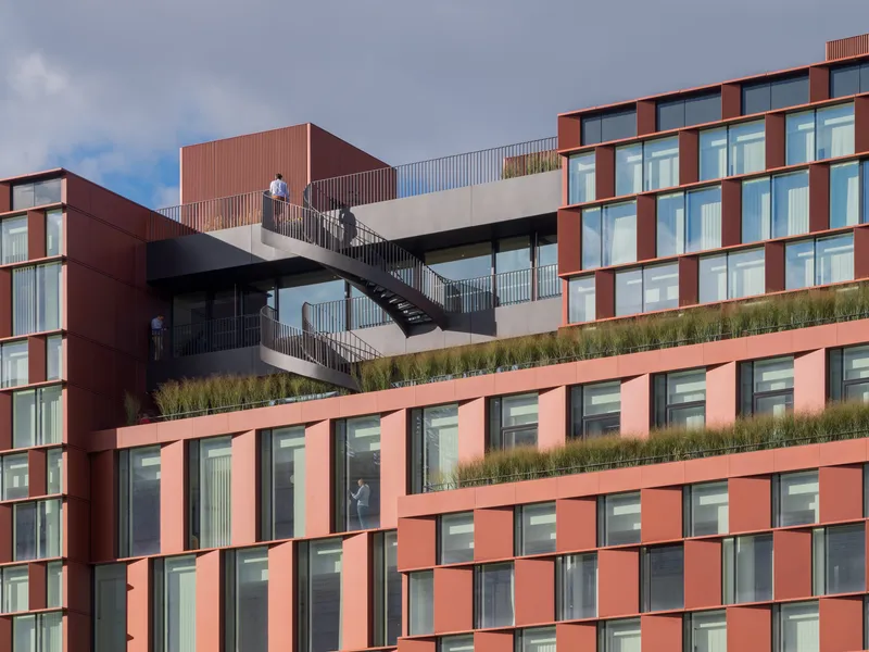 Exterior staircase with planted terraces connecting the red paneled facade levels under cloudy skies