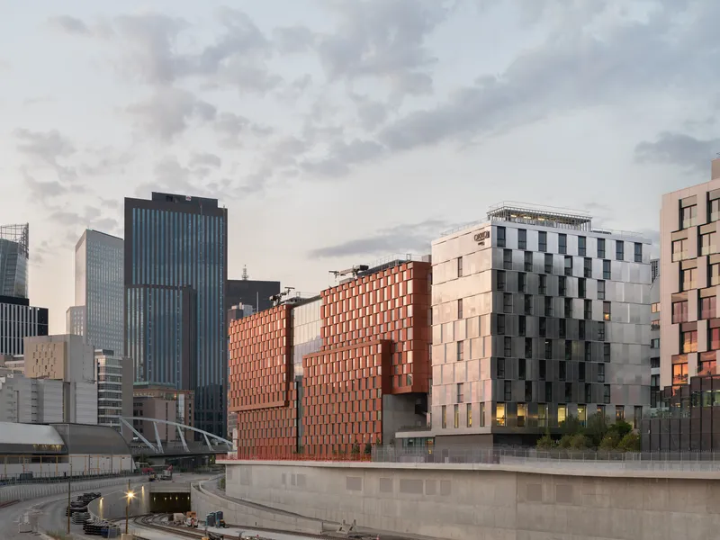 Urban context showing the red paneled volumes among neighboring towers at sunset beside a water feature