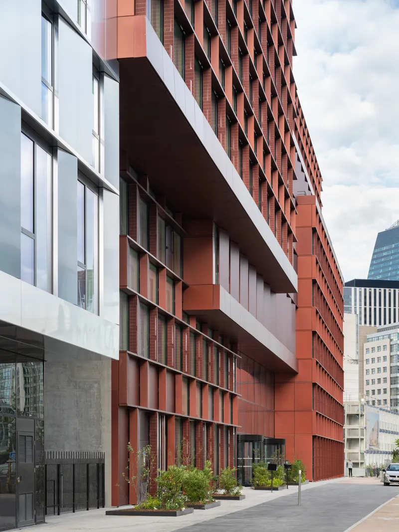Street-level view of stacked terracotta volumes above a concrete base with planted beds and pedestrians