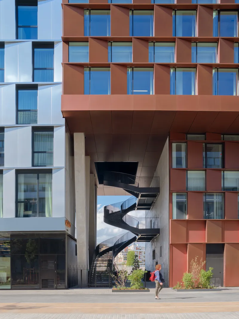 Spiraling black steel staircase between two residential volumes with a person walking below in sunlight