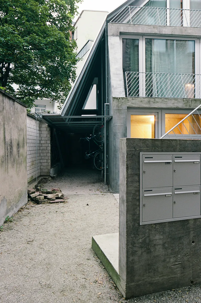 Gravel courtyard entry with black steel frame passageway and concrete mailbox beneath overhanging tree canopy