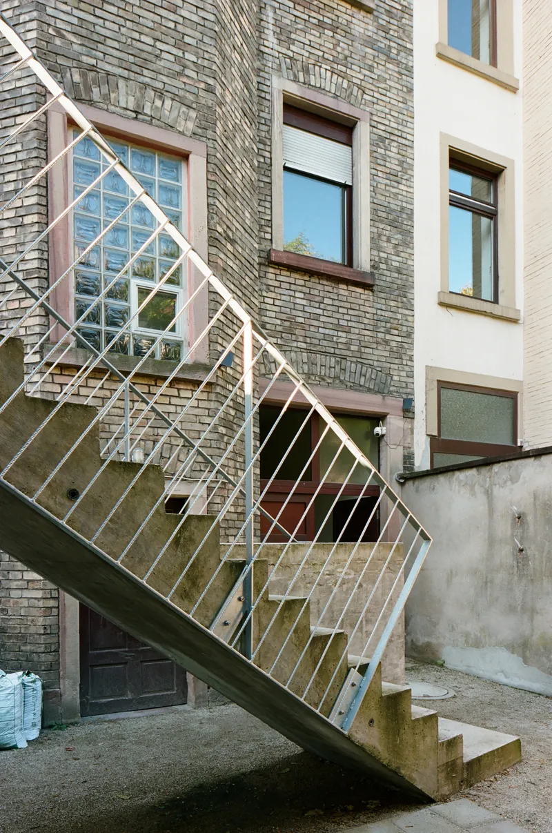 Diagonal view of steel staircase with white railings rising against grey brick facade with pink window frames