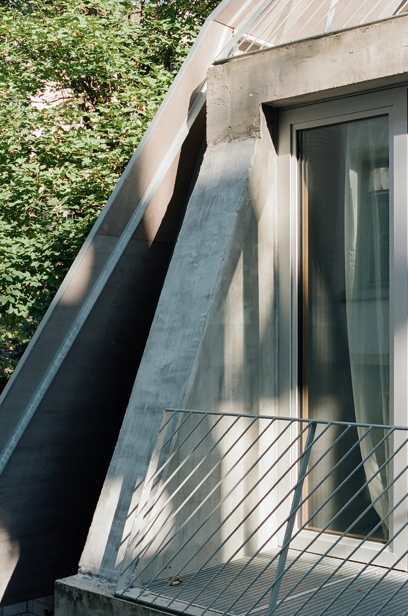 Angled facade detail with steel balcony railing and glazed door framed by concrete beams and foliage