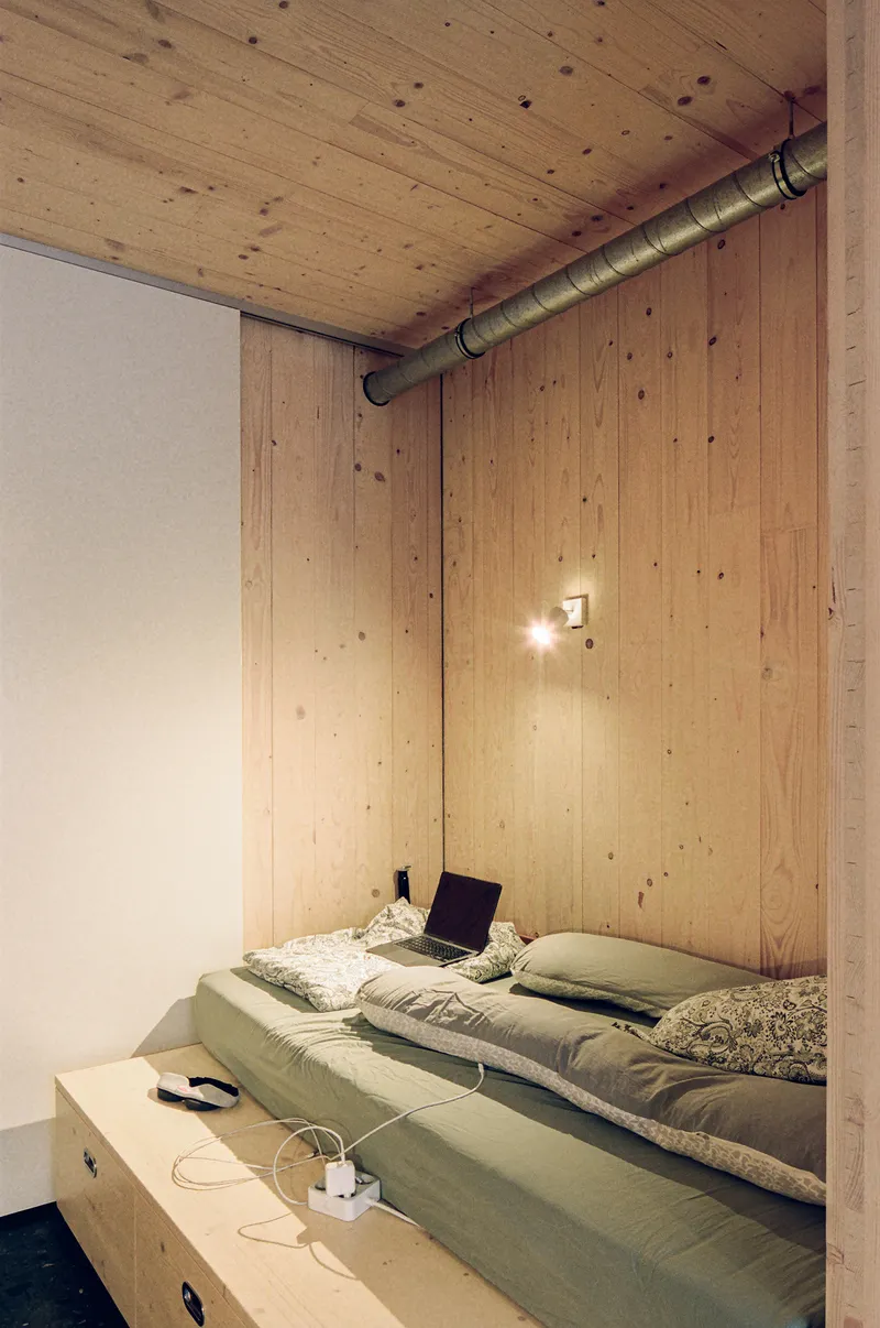 Sleeping platform with raised plywood bed base and exposed ventilation duct along the wood-clad wall