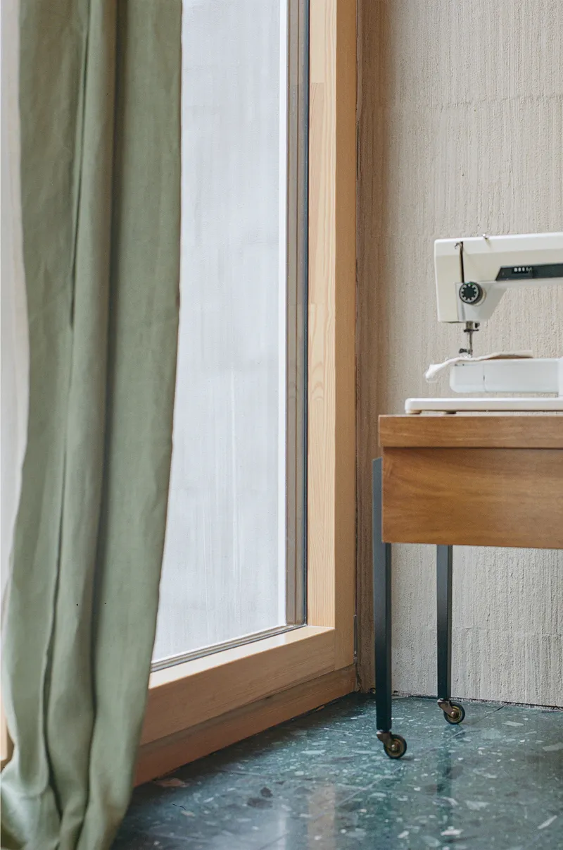 Timber window frame meeting mottled concrete floor with sewing table and sage green curtain nearby