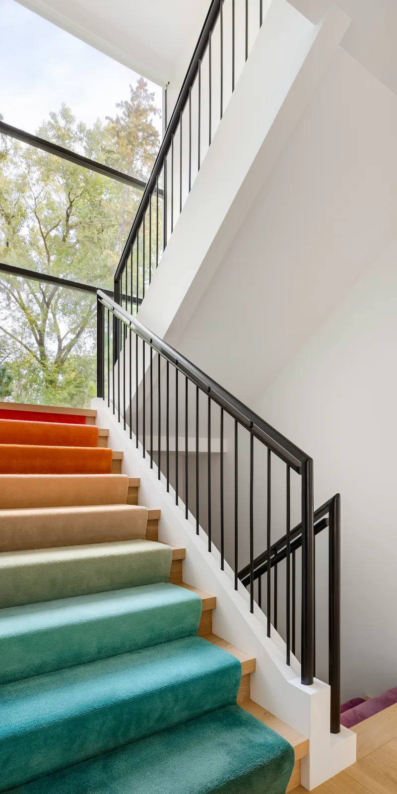 Staircase with gradient-colored runner from green to purple and black metal railing beside tall windows