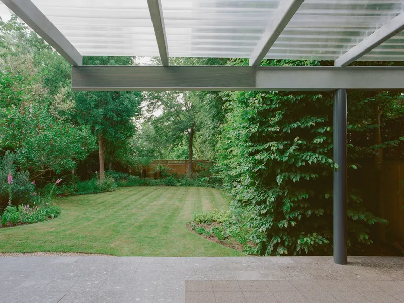 View from beneath the steel and polycarbonate canopy across the striped lawn toward wooded boundary