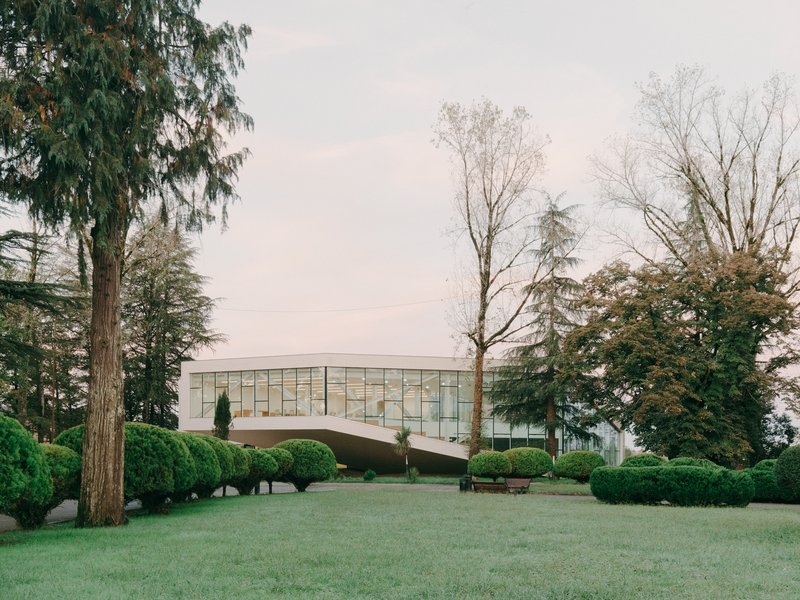 Cantilevered glazed facade with gridded window pattern surrounded by evergreen trees and clipped hedges on a lawn