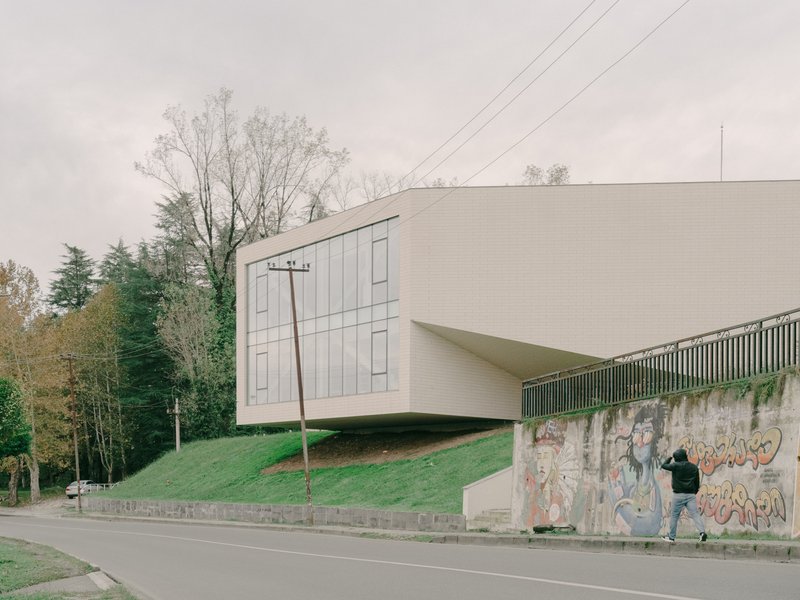 Street view of the elevated white volume above a graffitied retaining wall with a person walking below