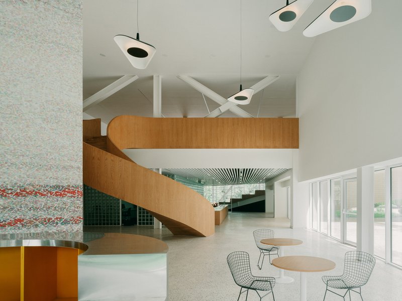 Double-height lobby with curving timber staircase, pendant lights, and wire mesh chairs at round tables