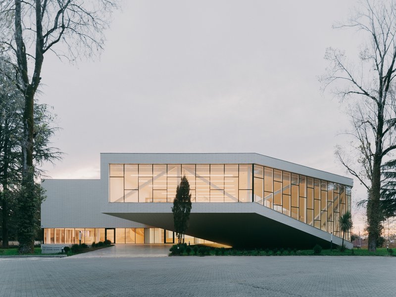 Elevated glazed volume supported by sloped walls among bare winter trees at dusk