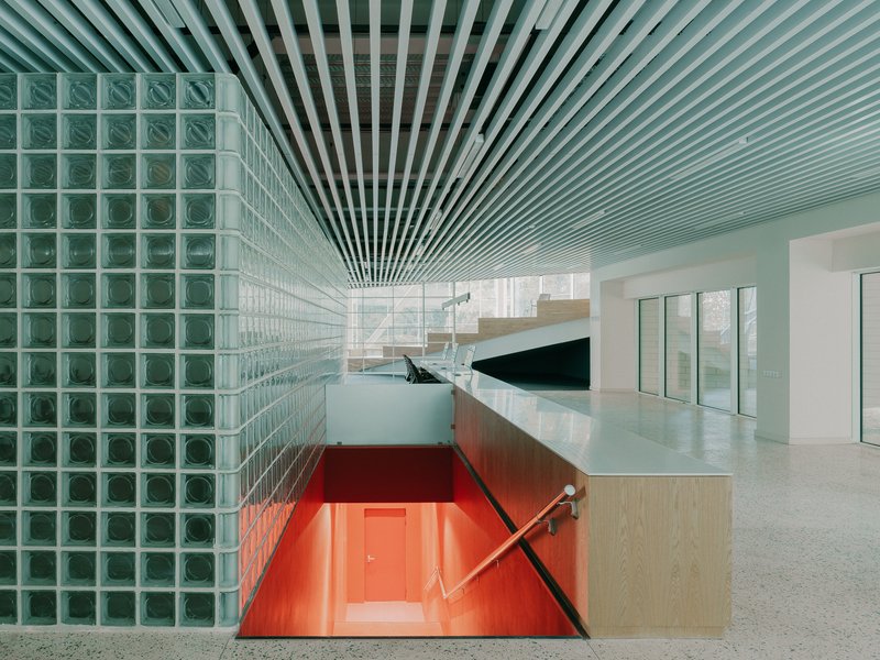 Interior view showing green glass block wall, radial slatted ceiling, and red-lit stairwell descending below
