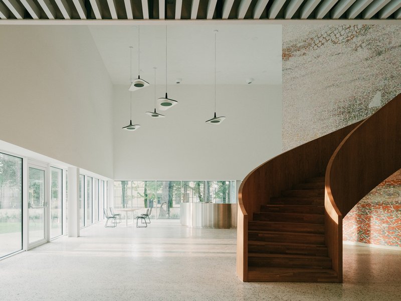 Curved timber staircase beside a textured wall with pendant lights overhead in a daylit lobby