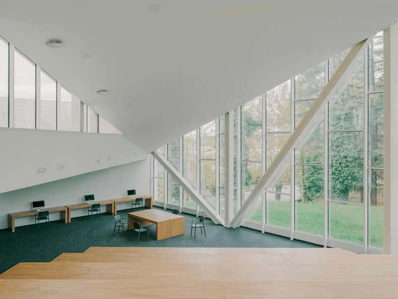 Double-height reading room with angled white structural beams framing floor-to-ceiling windows overlooking a park
