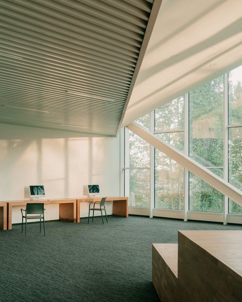 Study desks with green chairs under diagonal timber bracing against full-height windows in soft daylight