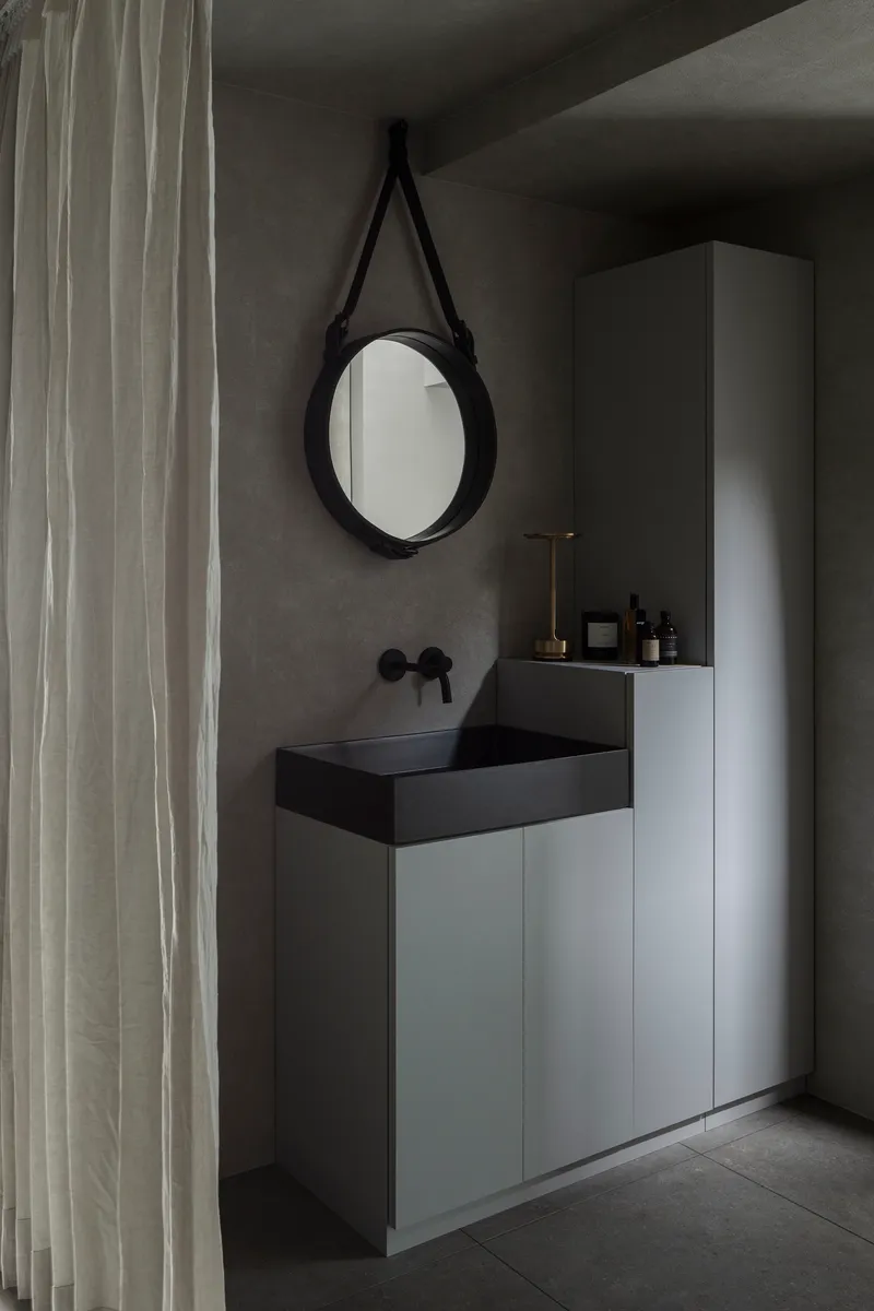 Bathroom vanity with black basin and round leather-strapped mirror beside linen curtain
