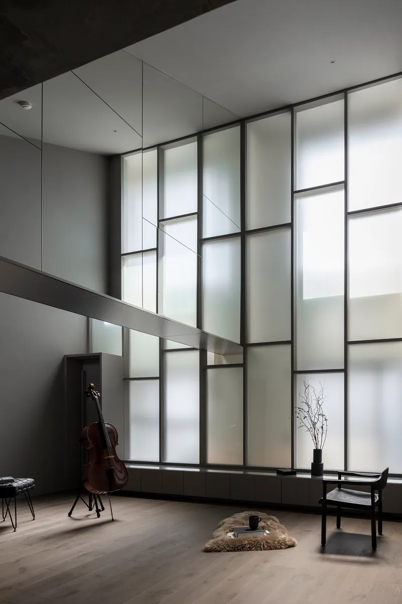 Double-height gridded glass wall illuminating gallery space with cello and minimal furnishings