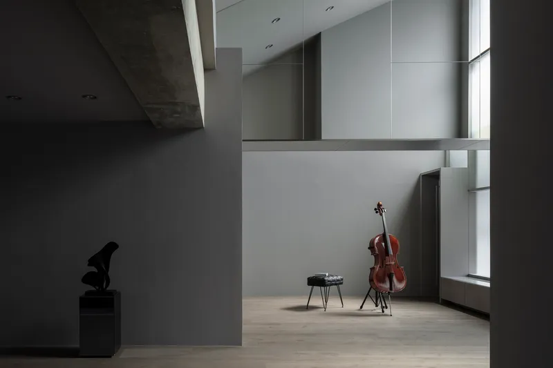 Gallery alcove with cello on stand beside narrow vertical window in grey walls