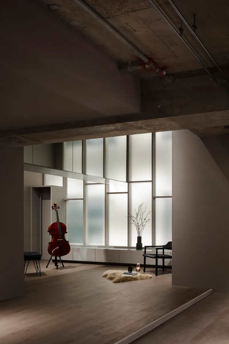 View through exposed concrete beam into space with gridded translucent glass partition and cello on stand