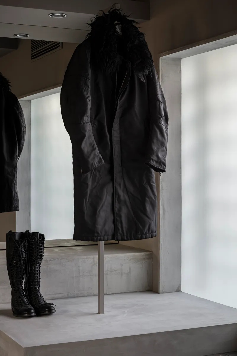 Close view of black coat suspended on metal stand above concrete plinth with boots