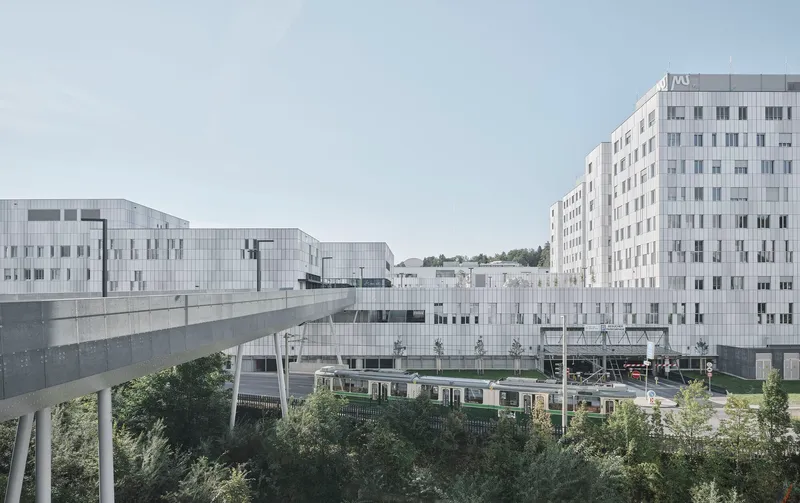 White metal panel facades connected by an elevated walkway above a passing train and green foliage